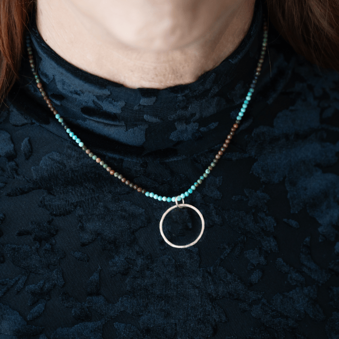 A woman wearing a turquoise beaded necklace with a hand-forged sterling silver open circle pendant at the collarbone, styled against a dark textured top.