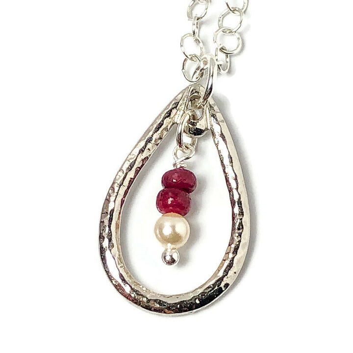Delicate hammered finish for incredible texture to give a vintage flair to the open teardrop pendant. Genuine Rubys-the queen of gems in a well-polished deep red and hand-cut rondelle shape. Swarovski crystal pearl to complement the ruby red of the gems. Lightweight pendant in rhodium-plated pewter. Handcrafted in CA. Pendant detail