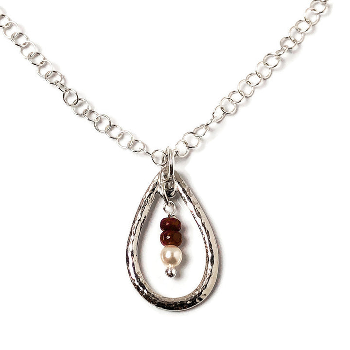 Delicate hammered finish for incredible texture to give a vintage flair to the open teardrop pendant. Genuine Rubys-the queen of gems in a well-polished deep red and hand-cut rondelle shape. Swarovski crystal pearl to complement the ruby red of the gems. Lightweight pendant in rhodium-plated pewter. Handcrafted in CA. Detail view